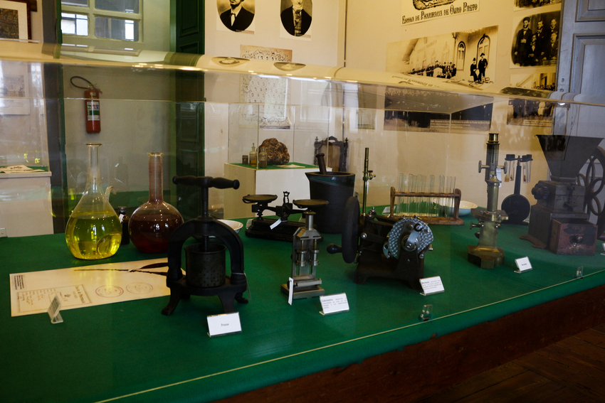 Vitrine de museu exibindo equipamentos e instrumentos científicos antigos, como frascos de vidro com líquidos coloridos, balanças, prensas e microscópios, sobre uma mesa verde com etiquetas de identificação.