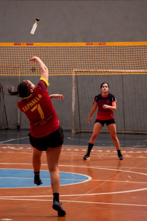 Atleta de peteca, com uniforme vermelho e o nome “Raphaela” nas costas, realiza um movimento para rebater a peteca durante a partida em quadra coberta. Outra jogadora observa atenta do lado oposto da rede.