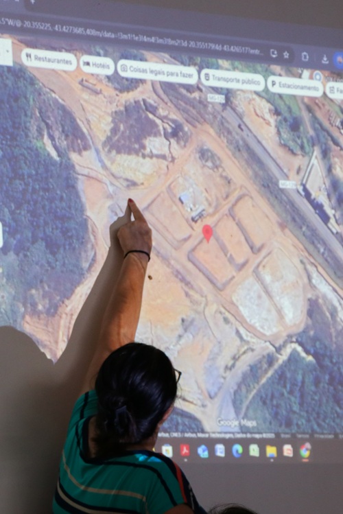 Pessoa aponta para uma projeção de imagem de satélite no Google Maps, mostrando uma área de terra com construções em andamento. A tela exibe área do Hospital Universitário.