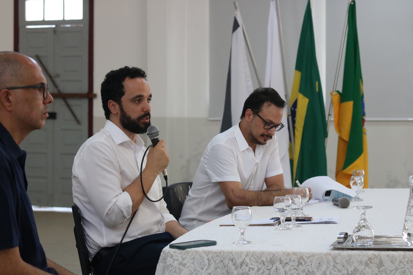 Três homens sentados em uma mesa de debate. O homem no centro, de barba e camisa branca, está falando ao microfone. Bandeiras do Brasil e outras estão ao fundo.