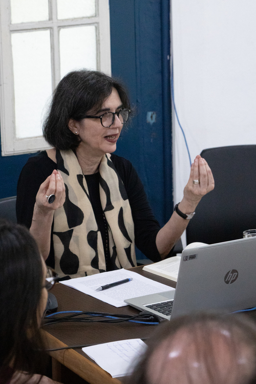 rofessora Doutora Cláudia Beltrão (Unirio) palestrando no XV Colóquio do LEIR/UFOP. Ela gesticula com as mãos enquanto fala em uma mesa, com um notebook HP e papéis à frente, vestindo uma blusa preta e um cachecol bege com estampa escura.