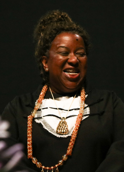 Mulher sorrindo em um ambiente interno, usando roupa escura com detalhes claros e colar longo ornamentado; seus cabelos estão presos e ela parece estar em um momento de alegria.