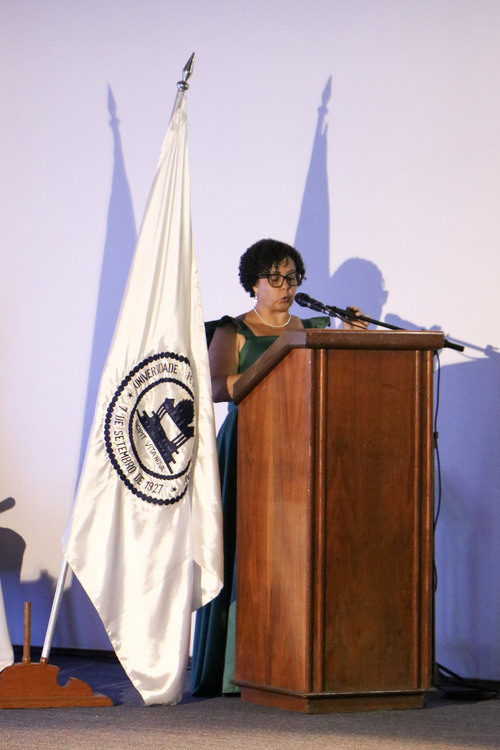 Pessoa de vestido verde está atrás de um púlpito de madeira, falando ao microfone e lendo um texto. Ao lado, uma bandeira institucional branca em suporte de chão. A iluminação azul sobre a parede ao fundo cria sombras marcadas da bandeira e da oradora.