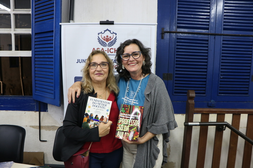 Duas mulheres sorrindo para a câmera, em frente a um painel de evento. As duas seguram livros em suas mãos.