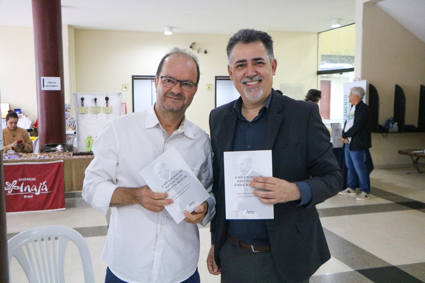 Dois homens posam sorridentes para uma foto em um ambiente interno, segurando livros acadêmicos. À esquerda, um homem de camisa branca e óculos; à direita, um homem de blazer preto e camisa escura. Ao fundo, há pessoas circulando e estandes de uma feira o