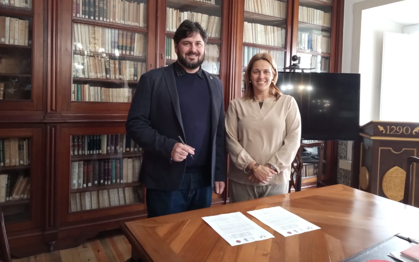 Imagem dos professores Felipe Comarela e Sandra Passinhas. À frente deles, há uma mesa com duas cópias do acordo assinados. 