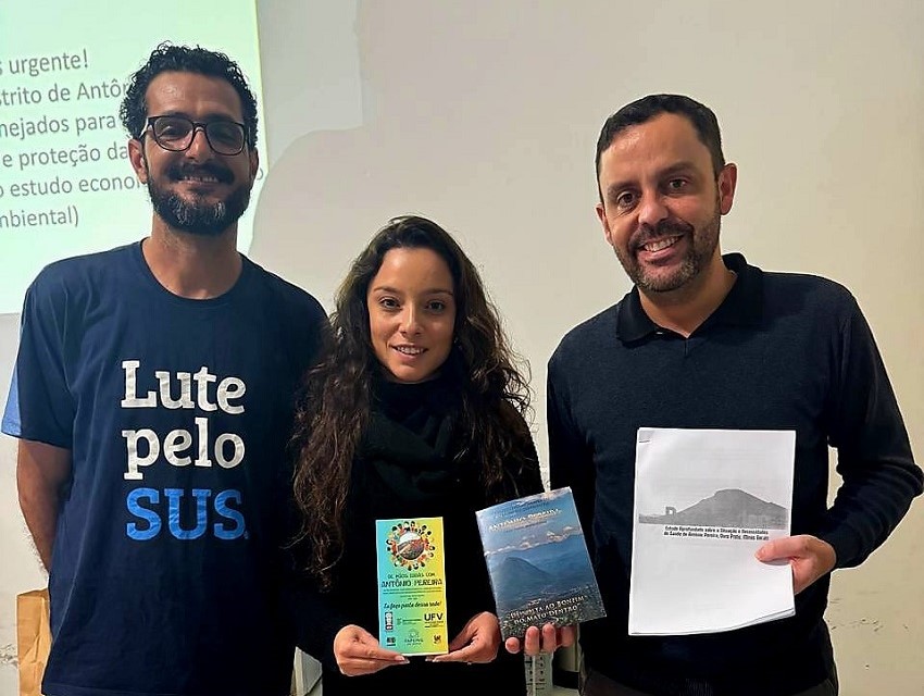 A imagem mostra três pessoas sorrindo em um evento. À esquerda, um homem usa uma camiseta com a inscrição "Lute pelo SUS". No centro, uma mulher segura um folheto, e à direita, um homem segura o relatório e um livro.