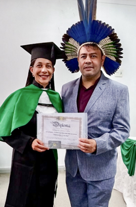 Professora sorridente em formatura, com beca verde e preta, ao lado de homem de terno azul e cocar indígena, segurando diploma.