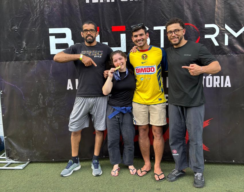 Na imagem, quatro pessoas posam sorridentes em frente a um banner de campeonato. A atleta ao centro exibe sua medalha, mordendo-a em sinal de conquista, enquanto os outros a abraçam e apontam para ela, celebrando juntos a vitória.