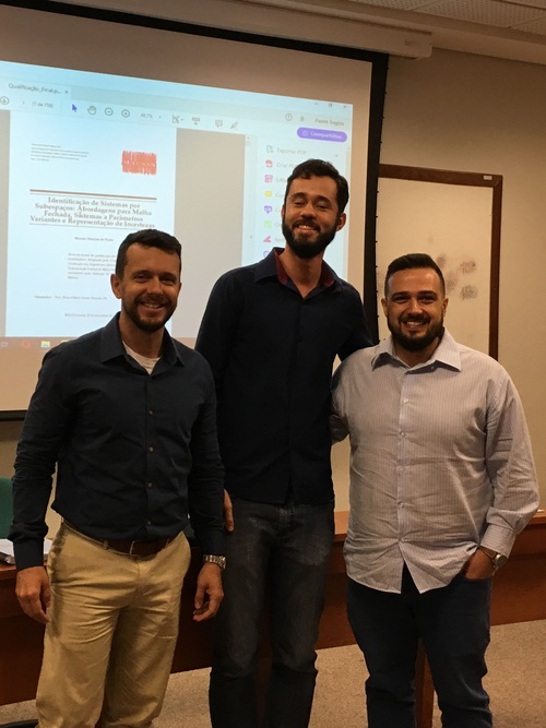 Três homens sorrindo em pé em frente a uma tela de projeção com um documento exibido, em ambiente formal, após defesa acadêmica. Todos usam camisas sociais e calças casuais.