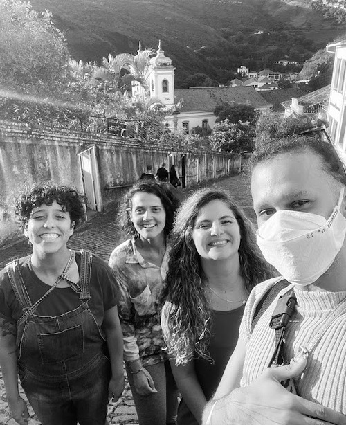 Foto em preto e branco de quatro pessoas sorrindo em uma rua de paralelepípedos com paisagem montanhosa ao fundo. Uma igreja colonial aparece ao longe. Uma das pessoas usa máscara facial.