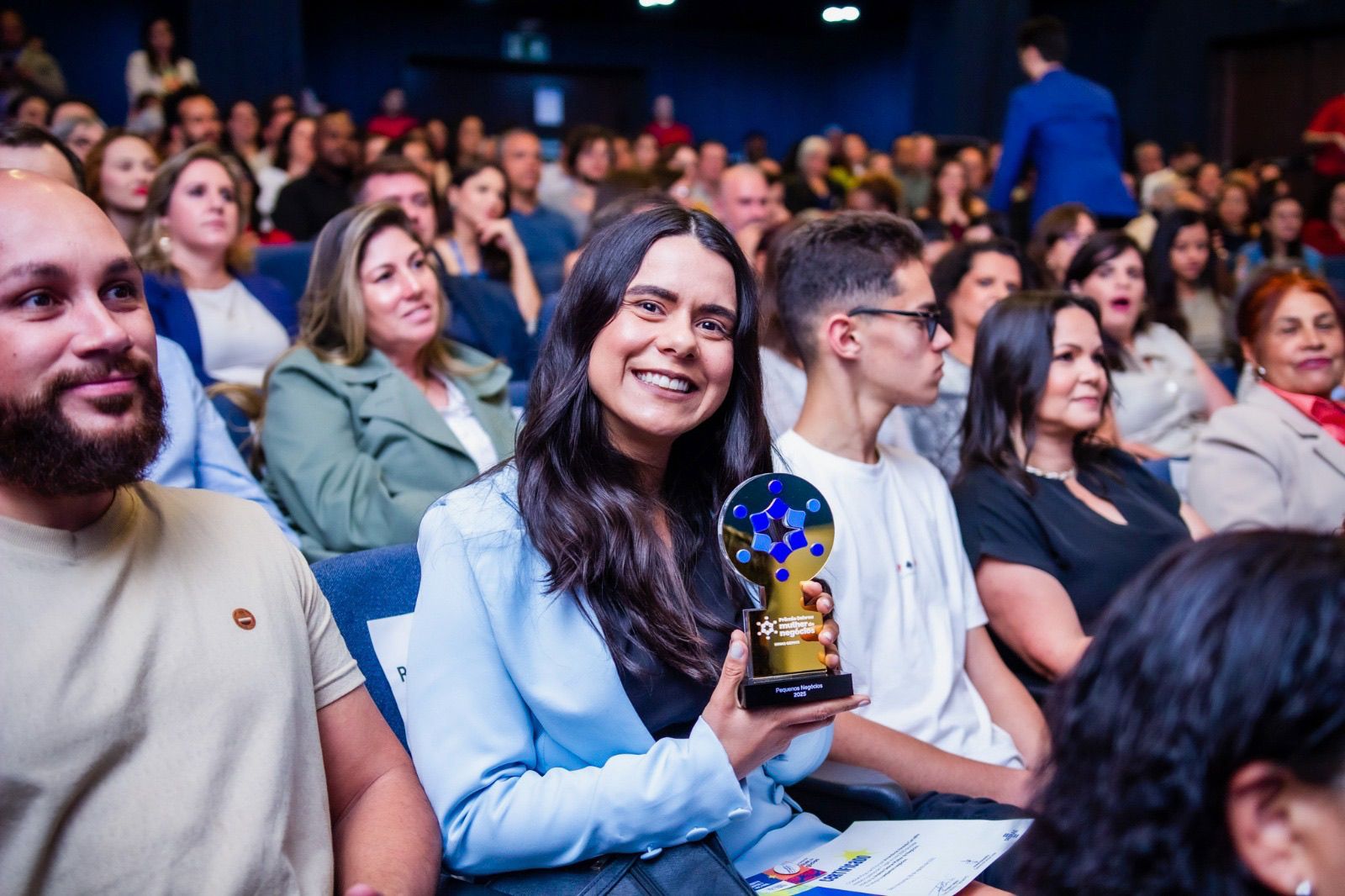 Imagem mostra Amanda Carvalho com o prêmio de Pequenos negócios na mão junto ao uma platéia. Amanda veste um blazer azul claro e posa para sorrido e com cabelos soltos.