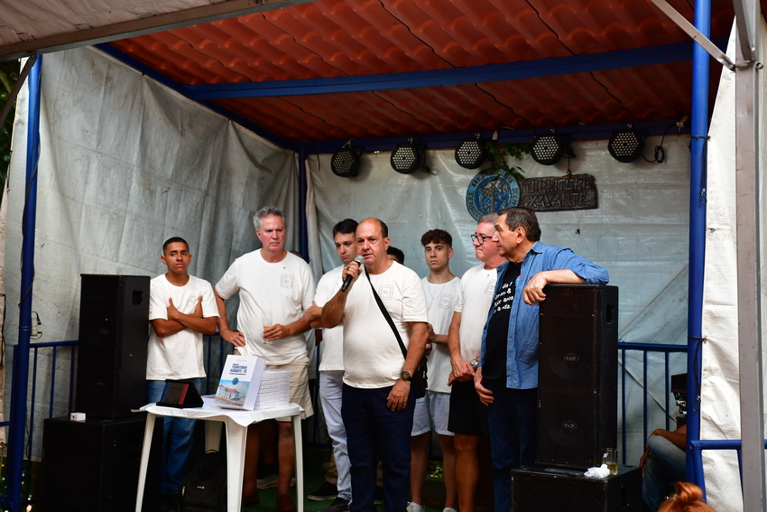 Fotografia de um grupo de homens reunidos em um pequeno palco coberto. Ao centro, um homem de camiseta branca fala ao microfone, enquanto os demais o acompanham. Há caixas de som, iluminação e uma mesa com livros expostos no palco.