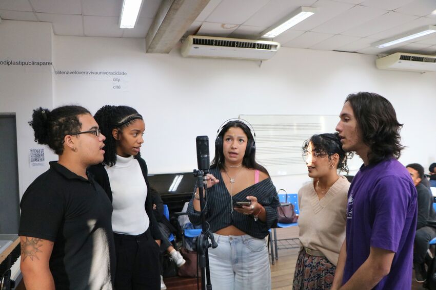 Cinco jovens estão reunidos em torno de um microfone em uma sala de aula.Duas pessoas usam roupas escuras, enquanto outras vestem peças em tons claros e roxo. Uma das participantes usa fones de ouvido e segura um celular nas mão. O grupo está concentrado 