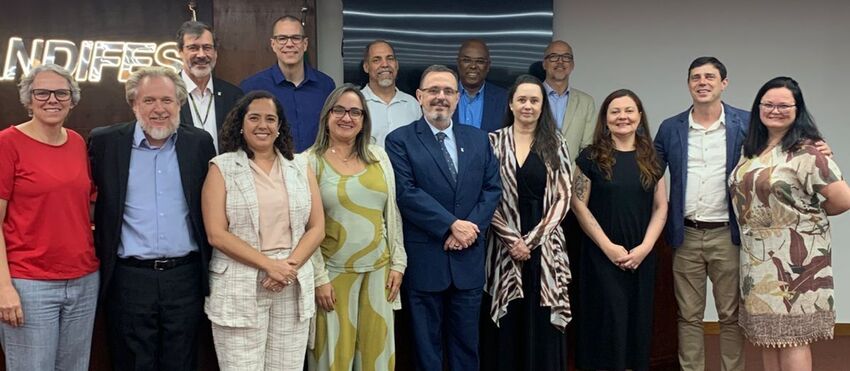 Treze pessoas, entre homens e mulheres, posam sorridentes em sala de reuniões iluminada, diante do letreiro “ANDIFES”. Vestem trajes sociais e semiformal, alinhados em uma única fileira. O encontro tratou da criação de uma comissão responsável pela organi