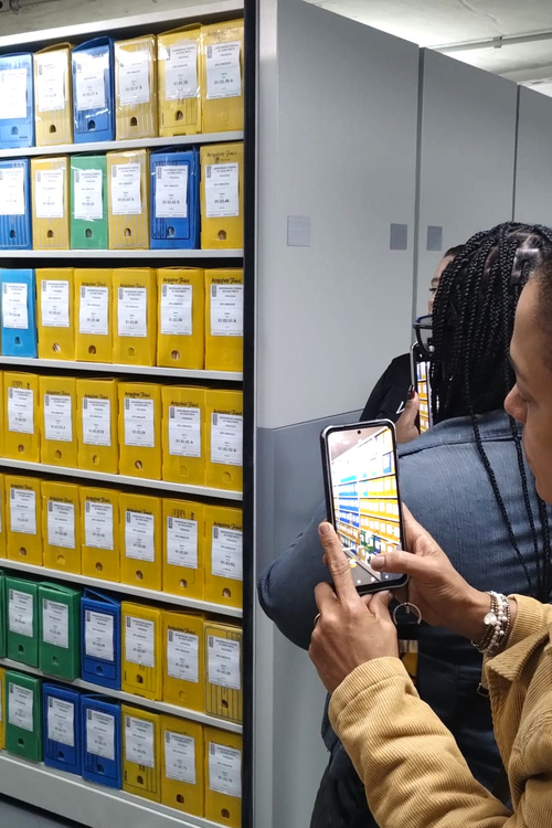 Pessoa tirando foto de uma estante de arquivos com pastas coloridas (azul, verde e amarela) organizadas em prateleiras. As pastas têm etiquetas visíveis, e uma das pessoas está segurando um celular para registrar a cena.