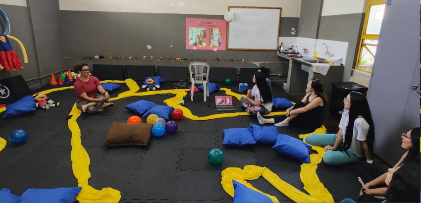 A foto mostra uma sala aberta e cheia de balões, almofadas, tecidos e brinquedos coloridos. Do lado direito, cinco mulheres estão sentadas. Elas estão olhando para uma mulher sentada à esquerda, que fala com elas.