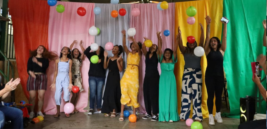 A foto mostra um grupo de 10 pessoas, com direntes cores, cabelos e corpos, comemorando, enquanto balões coloridos caem nelas. Elas estão em um palco e, nas laterais da foto, vemos braços e mãos das pessoas que assistem aplaudindo.