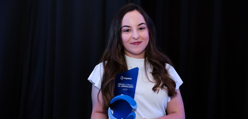 Carolina, uma jovem branca de cabelos escuros compridos, segura o prêmio, que é um troféu azul