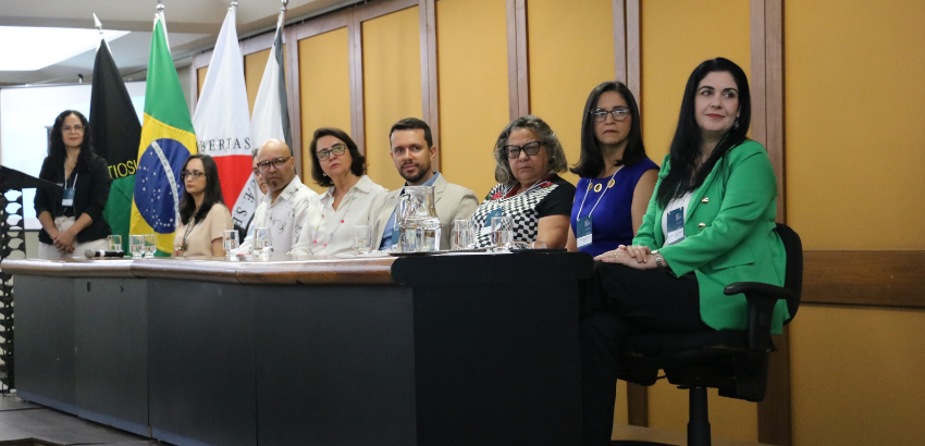 Foto aberta da mesa de abertura do VII CIM. Sete pessoas sentadas e uma em pé, no ponto mais distante, olham para a direita.