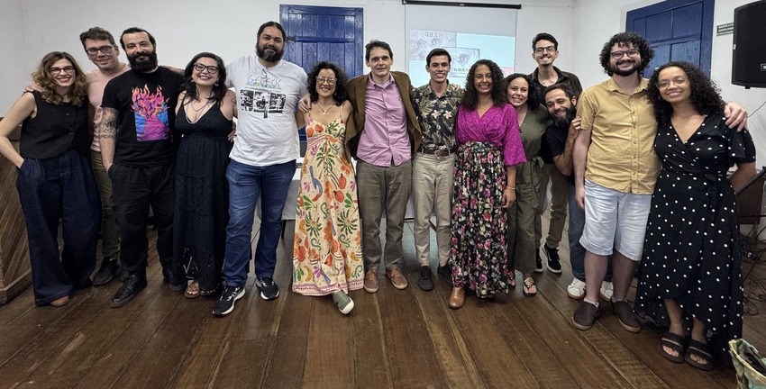Grupo de pessoas alinhadas lado a lado em uma sala, sorrindo para a câmera. Estão em pé sobre piso de madeira, com portas azuis ao fundo e um projetor ligado. A foto registra o grupo reunido no primeiro dia do evento.