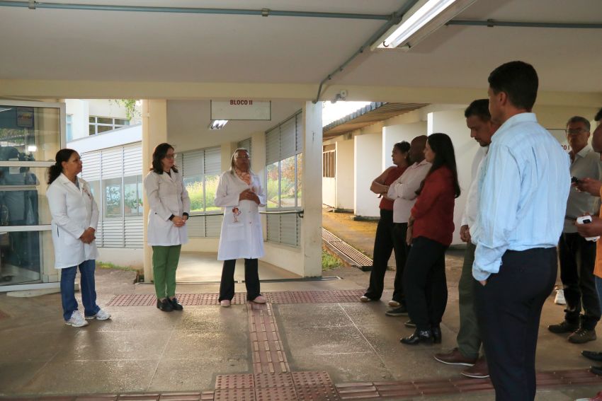 Em uma área coberta entre dois prédios, com piso tátil para acessibilidade, três mulheres de jaleco branco estão posicionadas à esquerda, voltadas para um grupo de pessoas à direita. Uma delas fala enquanto as outras observam.