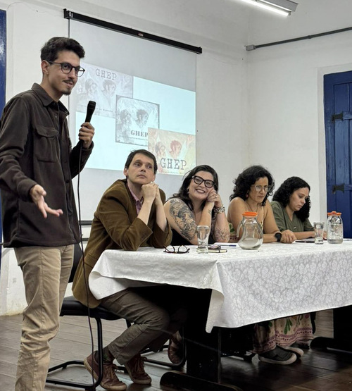 Homem em pé fala ao microfone ao lado de uma mesa onde quatro pessoas estão sentadas. Todos parecem participar de uma apresentação ou debate. Há jarros de vidro e papéis sobre a mesa, e um slide projetado ao fundo.