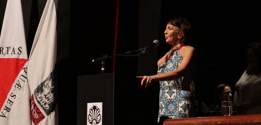 Adriana, uma mulher de aproximadamente 55 anos, é branca, usa um vestido colorido e fala ao púlpito.
