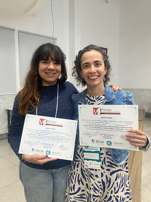 Duas mulheres sorriem para a câmera, segurando certificados de prêmio. A da esquerda tem cabelo castanho escuro e veste um suéter azul. A da direita, com cabelo cacheado escuro, usa uma jaqueta jeans sobre um vestido estampado. As duas usam crachás.
