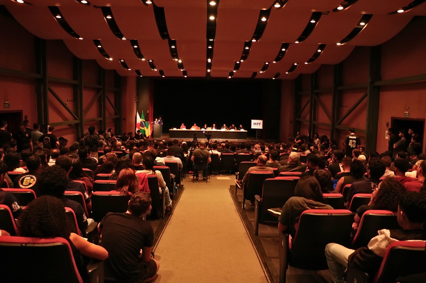 A imagem mostra uma audiência pública acontecendo em um auditório grande e moderno, com fileiras de poltronas vermelhas ocupadas por um público diverso e numeroso. A foto é tirada do fundo do auditório, de forma centralizada, oferecendo uma visão ampla.