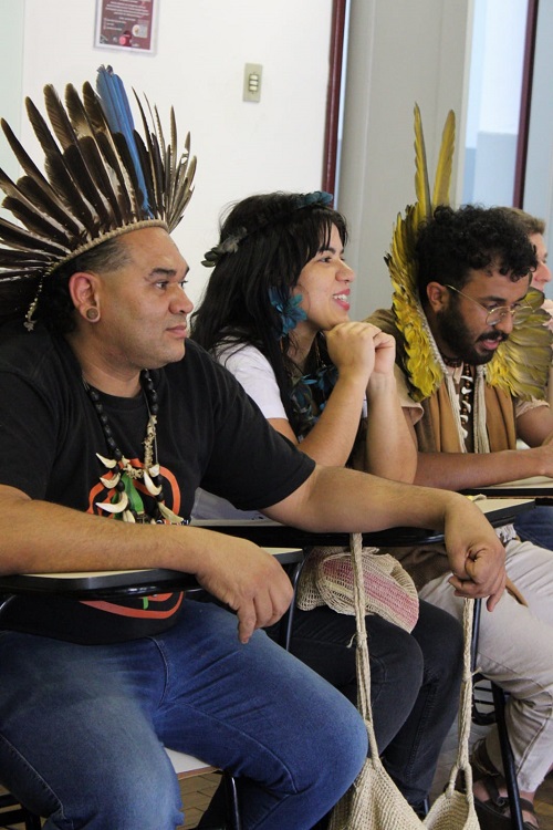 Três pessoas sentadas lado a lado em uma sala de aula participam de uma roda de conversa. Duas usam cocares e colares tradicionais, enquanto a pessoa ao centro sorri. Há carteiras escolares e bolsas de tecido penduradas.