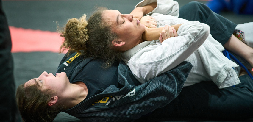 Na imagem, duas atletas de Jiu-Jitsu estão em combate. A lutadora de kimono branco aplica um estrangulamento pelas costas, enquanto a oponente, de kimono preto, tenta resistir à pressão. A cena mostra concentração e intensidade no tatame.