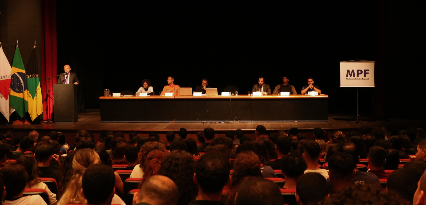  No palco, há uma mesa comprida com sete pessoas sentadas, todas voltadas para o público. À esquerda da mesa, um homem está de pé em um púlpito, falando ao microfone. 