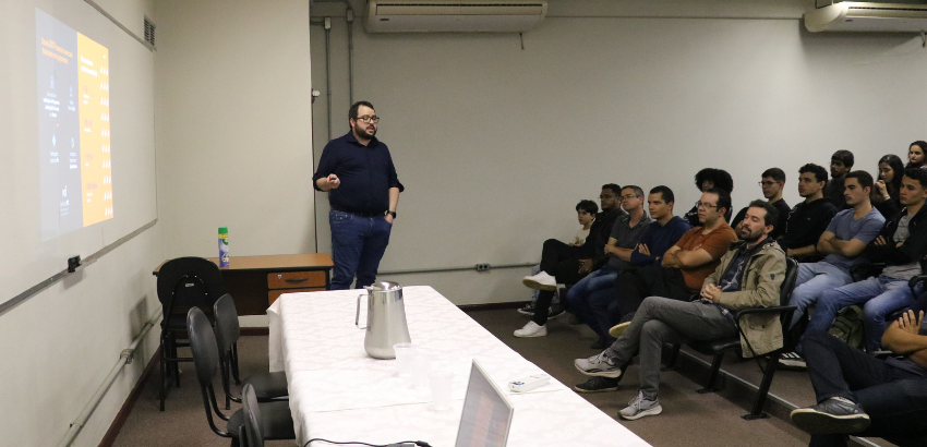 A imagem mostra o palestrante Denis falando aos estudantes na plateia. 