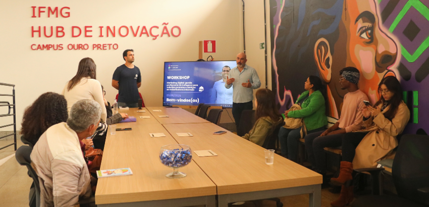 A imagem mostra um grupo reunido em torno de uma mesa de reuniões, enquanto um palestrante apresenta informações em uma tela digital. Ao fundo, uma parede com arte urbana colorida e os dizeres "IFMG Hub de Inovação Campus Ouro Preto". 