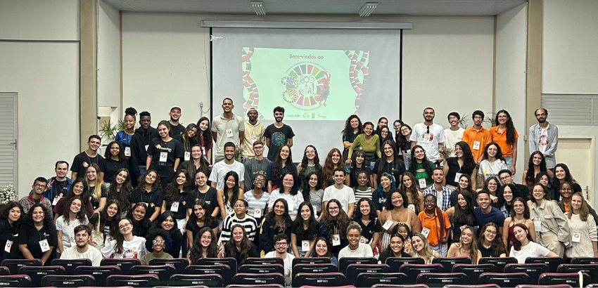 Foto de um grande grupo de pessoas reunidas em um auditório. Todos estão posando para a foto sorrindo, com um logotipo colorido abaixo do evento. Há cerca de 80 pessoas, organizadas em várias fileiras: algumas em pé no palco, outras sentadas ou ajoelhadas