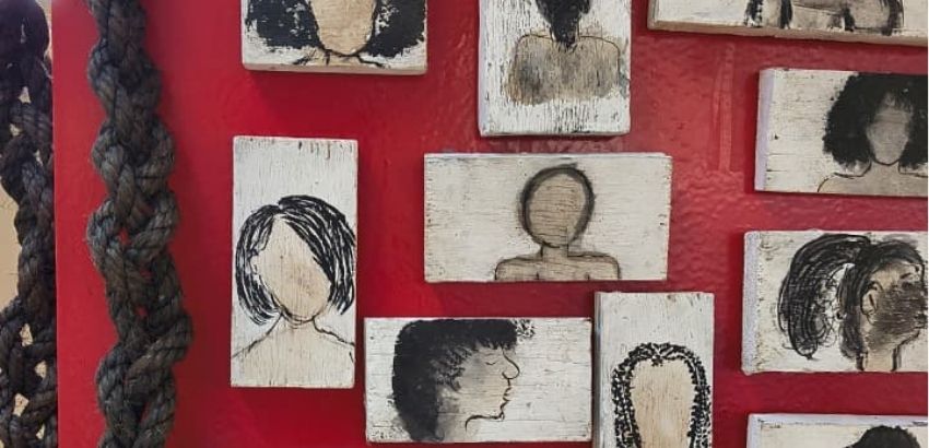 Painéis com ilustrações de diferentes penteados e silhuetas femininas em madeira clara estão expostos sobre uma parede vermelha. Ao lado esquerdo da imagem, grandes tranças pendem verticalmente, compondo a instalação artística.