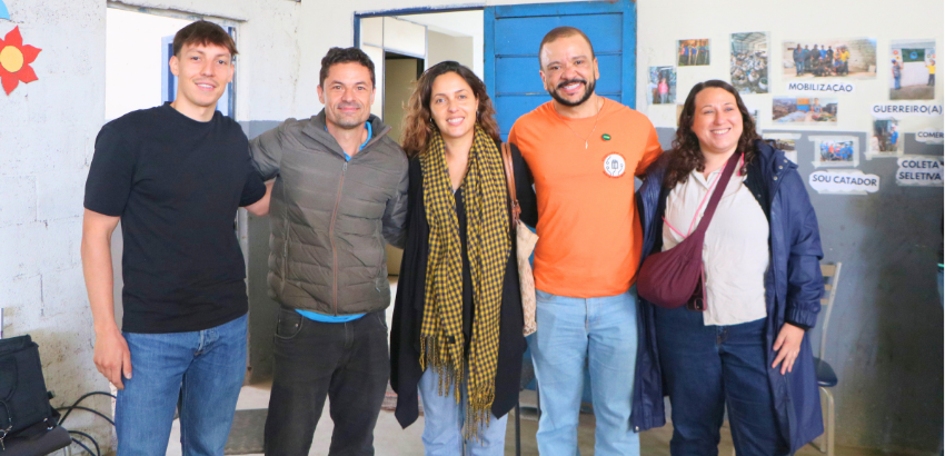 Cinco pessoas posam lado a lado para uma fotografia em um ambiente interno, sorrindo para a câmera. Da esquerda para a direita, há um homem de camiseta preta, um homem com jaqueta verde, uma mulher com cachecol estampado, um homem de camiseta laranja e um