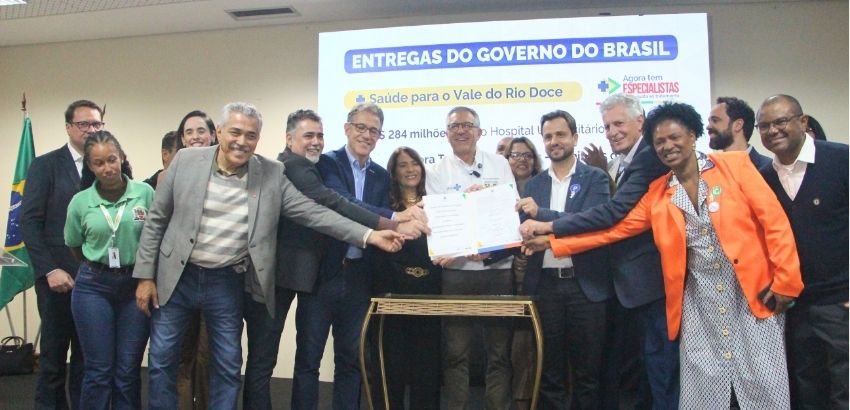 Fotografia de uma cerimônia institucional realizada em ambiente interno. Cerca de treze pessoas estão reunidas em frente a uma tela de projeção que exibe o título “Entregas do Governo do Brasil” e informações sobre investimentos em saúde para o Vale do Ri