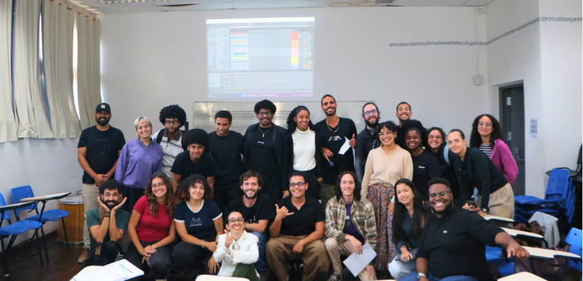 Fotografia de grupo tirada em uma sala de aula universitária. O grupo é majoritariamente composto por jovens adultos, muitos sorrindo e fazendo gestos descontraídos. Ao fundo, há uma projeção ligada em uma parede branca, exibindo a interface de um softwar