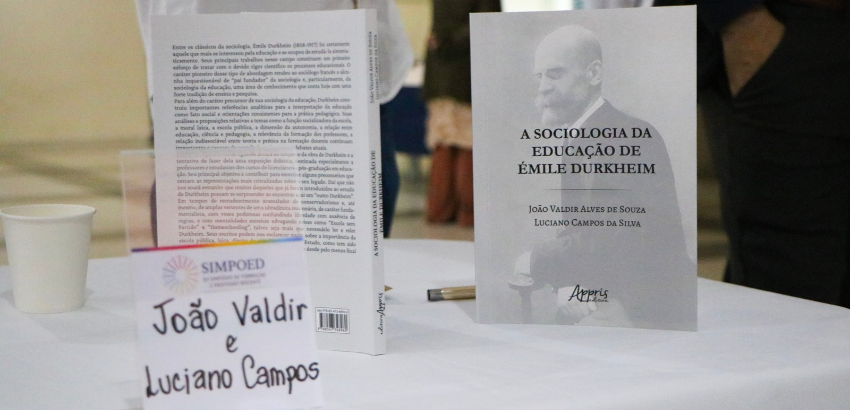 Livro “A Sociologia da Educação de Émile Durkheim”, de autoria de João Valdir Alves de Souza e Luciano Campos da Silva, exposto sobre uma mesa durante um evento acadêmico. Ao lado, há um exemplar aberto mostrando o conteúdo interno da obra e um cartaz com