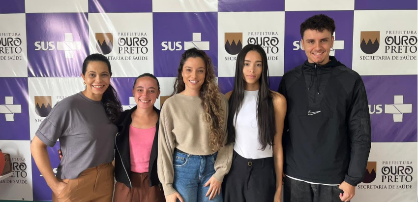 Cinco pessoas posam lado a lado em frente a um painel quadriculado nas cores roxa e branca, com logotipos da Secretaria de Saúde e da Prefeitura de Ouro Preto. Da esquerda para a direita, há uma mulher de camiseta cinza e calça marrom, uma mulher de blusa