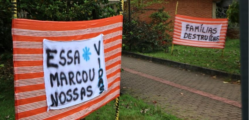Faixas de tecido listradas em vermelho e branco estão expostas em uma área arborizada, com cartazes escritos à mão com as frases “Essa marca nossas vidas” e “Famílias destruídas”.