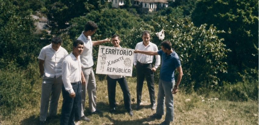 Fotografia de seis homens em uma área gramada, cercada por vegetação, segurando e apontando para uma placa com os dizeres “Território Avante República”. Ao fundo, há árvores, arbustos e algumas construções.