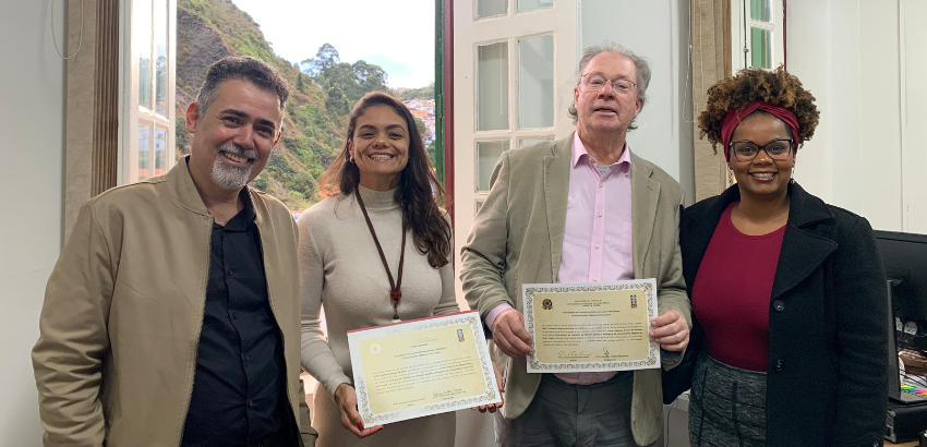 Quatro pessoas posam sorrindo para a foto em um ambiente interno, possivelmente uma sala administrativa com janelas abertas ao fundo, revelando uma paisagem com morros e casas. Duas pessoas ao centro seguram certificados.