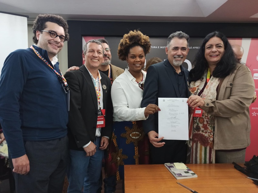 Grupo de sete pessoas sorri para a câmera durante evento, segurando um documento oficial assinado. Ao fundo, há um painel com faixas vermelhas. Todos usam crachás, indicando participação institucional.          Perguntar ao ChatGPT