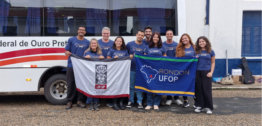 Foto de dez pessoas participantes do projeto segurando duas bandeiras, uma da UFOP e outra do Projeto Rondon, em frente a um ônibus