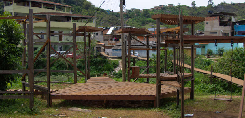 Foto de plano geral da praça-teatro, construída de madeira, com um balançador na esquerda e uma parte central espaçosa, na imagem possui casas e a ponte ao fundo, com mato em volta da estrutura