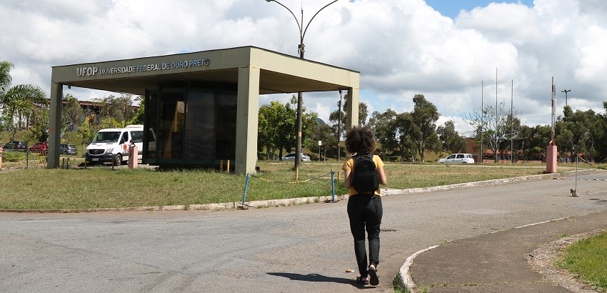 Vista da entrada da Universidade Federal de Ouro Preto com um ponto de ônibus e uma pessoa caminhando em direção a ele, em um dia claro