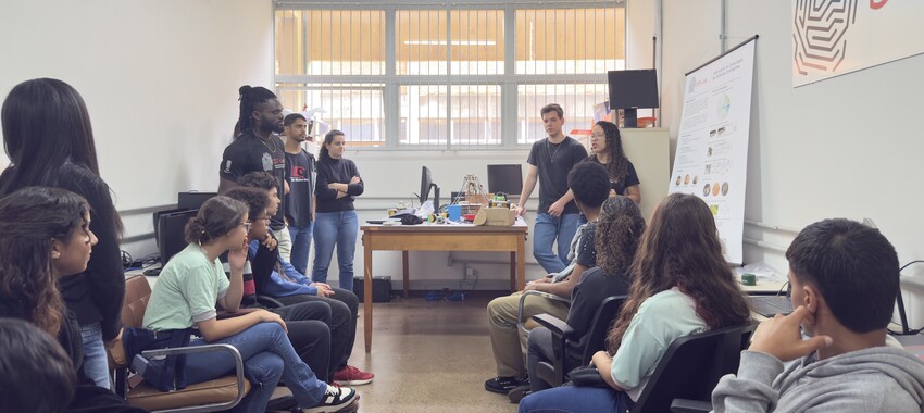 Alunos e instrutores em uma sala de aula clara, com grandes janelas, participam de uma apresentação ou oficina. Quatro pessoas estão de pé perto de uma mesa com equipamentos, enquanto um grupo maior está sentado em cadeiras.